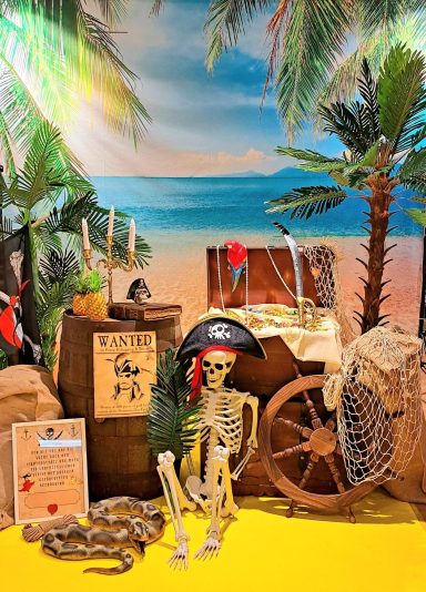 Skelett in Piratenkostüm vor einer Schatzkiste und tropischer Kulisse.