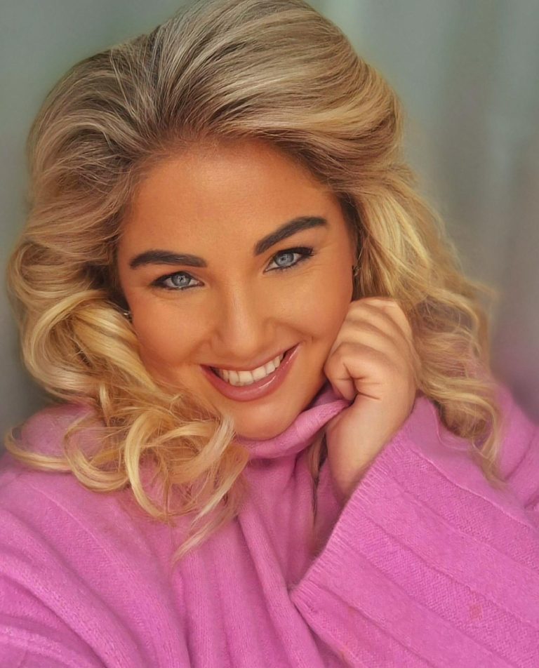 Lächelnde Frau mit blonden Locken und blauen Augen in einem pinken Pullover.