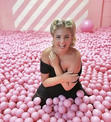 Frau sitzt lächelnd in einem Meer aus rosa Bällen mit lila und pinken Hintergründen.