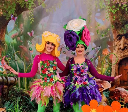 Zwei Frauen in bunten Blumenkostümen stehen in einer fantasievollen Gartenlandschaft.